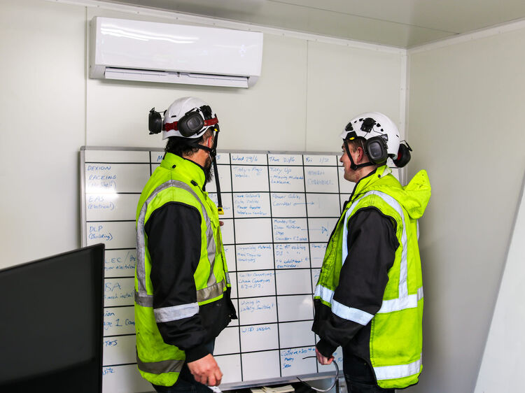 Two technicians in hi-vis and hard hats reviewing a job board in a site office
